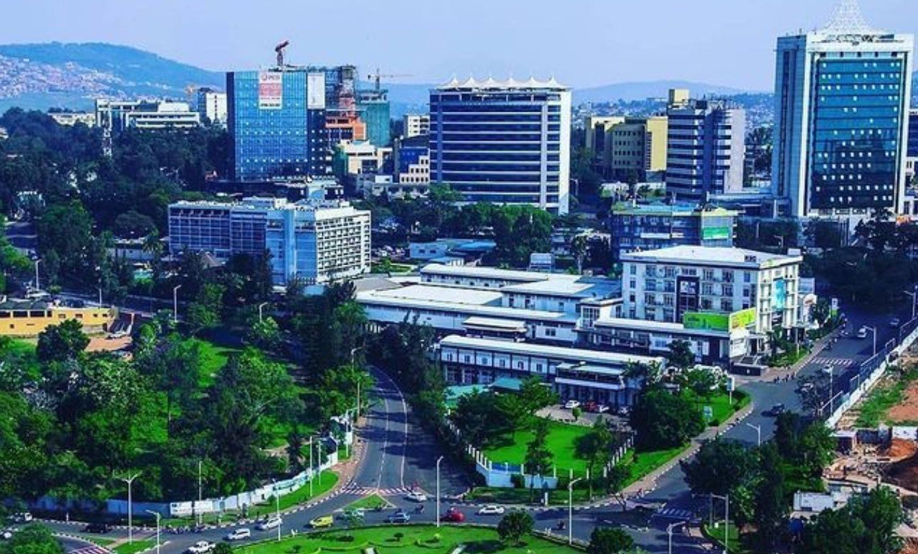 Aerial view of a modern city skyline with office buildings, green parks, roads, and mountains in the background under blue sky